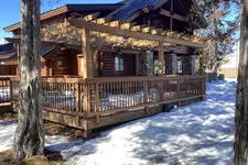 Thumbnail for Wooden log cabin with stone chimney surrounded by snow and tall trees under clear blue sky.