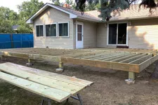 Thumbnail for Wooden deck frame under construction attached to beige house with windows and door in backyard.