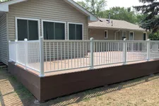Thumbnail for Spacious elevated wooden deck with white railing attached to beige house surrounded by greenery on a sunny day