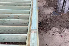 Thumbnail for Close-up of wooden deck framing with beams and joists under construction near tree trunks and dirt ground.