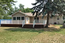 Thumbnail for Single-story beige house with a large wooden deck, white railing, and green lawn under a tall tree.