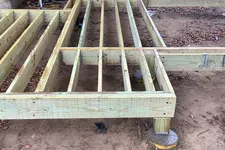 Thumbnail for Wooden deck frame construction with metal supports next to a log cabin on a dirt ground.