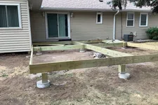 Thumbnail for Wooden deck foundation frame built on concrete piers next to a beige house with sliding glass door.