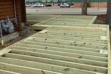 Thumbnail for Close-up of wooden deck framing attached to log cabin exterior under cloudy sky with street and buildings in background