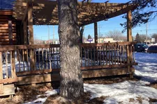 Thumbnail for Wooden pergola and deck around a tree with patches of snow and dry grass under a clear blue sky.