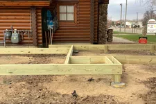 Thumbnail for Wooden frame construction beside a log cabin with stone accents and metal roof in an outdoor setting near a street.