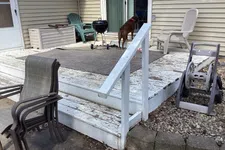 Thumbnail for Outdoor wooden deck with peeling white paint, patio chairs, a grill, a dog looking inside sliding glass door.