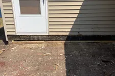 Thumbnail for Sunlit area in front of a house with beige siding, a white door, and a brick ground partially covered in debris.