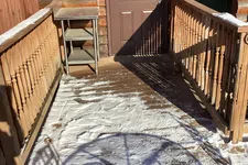 Thumbnail for Snow-covered wooden porch with boot prints, railing shadows, and a closed brown door on a log cabin.