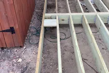 Thumbnail for Wooden deck frame construction next to a red shed and log cabin wall on rocky soil ground.