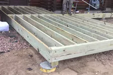 Thumbnail for Construction worker building wooden deck frame outside a log cabin on a dirt and gravel surface.
