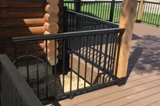 Thumbnail for Brown wooden deck with black metal railing and part of a log cabin under clear daylight.