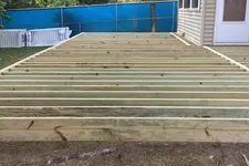 Thumbnail for Construction of a wooden deck frame attached to a house in a backyard with trees and a blue fence.