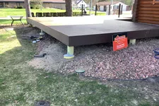 Thumbnail for Raised wooden deck platform supported by posts over gravel and grass near a log cabin exterior.