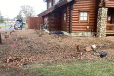 Thumbnail for Prepared ground with stakes and markings next to a log cabin under construction with stone pillar details.