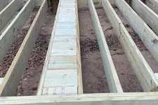 Thumbnail for Wooden floor joists and beams laid out for deck construction next to a log cabin exterior door.