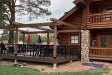 Thumbnail for Outdoor wooden deck with black railing and pergola attached to a rustic log cabin surrounded by green grass.