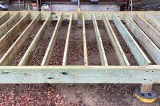 Thumbnail for Wooden deck frame under construction in front of a log cabin with soil and gravel ground.