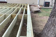 Thumbnail for Partially constructed wooden deck frame next to a tree outside a house with beige siding.