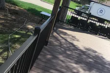 Thumbnail for Spacious backyard deck with brown wooden flooring, black metal railing, and a white truck parked nearby on a sunny day.