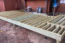 Thumbnail for Wooden deck frame construction beside a log cabin on dirt ground, with tools and gas meters nearby.