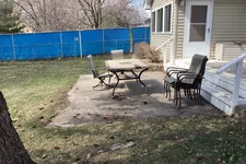 Thumbnail for Backyard patio with outdoor table, four chairs, and house stairs under clear sky, fenced by blue mesh screen.