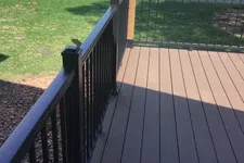Thumbnail for Outdoor deck with dark brown wooden floorboards and black metal railing overlooking a road and green lawn.