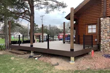 Thumbnail for Partially constructed wooden deck attached to a log cabin with black railings and vertical posts on a cloudy day.