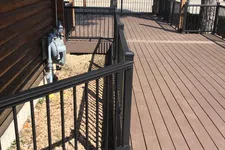 Thumbnail for Spacious brown wooden deck with black metal railings next to a log cabin and trees in the background.