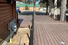 Thumbnail for Wooden deck adjacent to a log cabin with metal railing, trees, broom, and parked cars in the background.