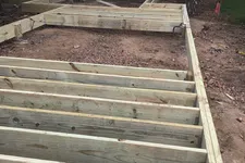 Thumbnail for Wooden deck frame under construction on dirt ground with nearby trees and road in the background.