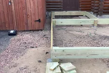 Thumbnail for Construction of wooden deck frame beside log cabin with stacks of lumber and gravel ground