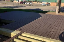 Thumbnail for Partially completed wooden deck with exposed beams in foreground and street with buildings and parked cars in background.