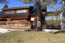 Thumbnail for Wooden log cabin with black metal roof, large porch, trees, and patches of snow on the ground under clear blue sky