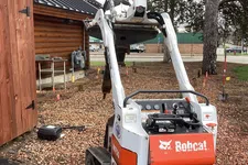 Thumbnail for Compact Bobcat MT55 mini track loader parked near a wooden cabin on a construction site.