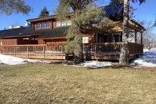 Thumbnail for Rustic wooden cabin with black metal roof, large deck, surrounded by snow patches and trees on sunny day.