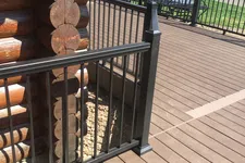 Thumbnail for Brown wooden deck with black metal railing attached to a log cabin corner, outdoor suburban setting.
