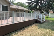 Thumbnail for Wooden backyard deck with white railing and stairs attached to a beige house, surrounded by grass and trees.