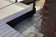 Thumbnail for Close-up of brown composite decking and railing next to a log cabin corner with exposed ground and cables.