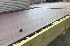 Thumbnail for Partially installed dark composite decking boards on a wooden deck frame next to a house exterior in daylight.