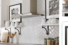 Thumbnail for White kitchen with herringbone tile backsplash, stainless steel hood, open shelves with white ceramics, and glass jars.