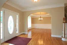 Thumbnail for Bright empty living room with hardwood floors, beige walls, white trim, oval glass front door, and chandelier.
