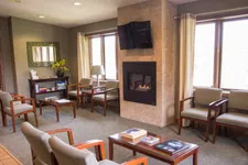 Thumbnail for Cozy waiting room with cushioned chairs, a fireplace, flat-screen TV, and natural light from large windows.