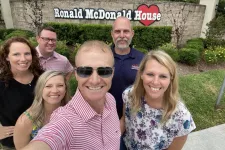 Thumbnail for Group selfie of six smiling adults in front of Ronald McDonald House sign outdoors on a sunny day