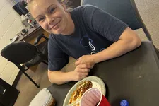 Thumbnail for Young woman smiling at a table with a plate of food, a red cup, and a soda bottle in an office setting.
