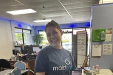 Thumbnail for Smiling woman wearing a MaidPro shirt stands in a bright office with chairs, a table, and documents.