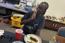 Thumbnail for Person wearing MaidPro shirt making peace sign while sitting at table with food and drink in break room.