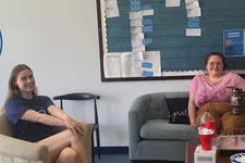 Thumbnail for Two women sitting in a casual office lounge with a bulletin board and coffee table with snacks.