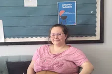 Thumbnail for Smiling woman in pink shirt and brown pants sitting cross-legged on light gray couch with bulletin board background.