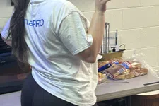 Thumbnail for Woman in glasses and white T-shirt posing in a kitchen area with food items and cleaning supplies visible.